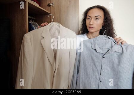 Un giovane uomo serio che guarda lo specchio e pensa se la camicia grigia sarà grande con la giacca crema Foto Stock