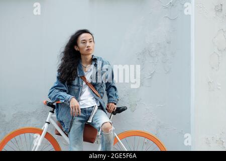 Elegante giovane in jeans e giacca in denim che pende in bicicletta e guarda lontano Foto Stock
