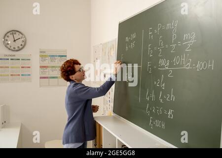 Insegnante grazioso con il pezzo di formula di scrittura del gesso sulla lavagna alla lezione della fisica Foto Stock