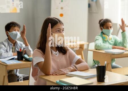 Gruppo di scolari in maschere protettive che alzano le mani alla lezione Foto Stock