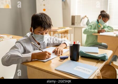 Diligente scolaro asiatico in maschera protettiva di scrittura in copybook contro il compagno di classe Foto Stock