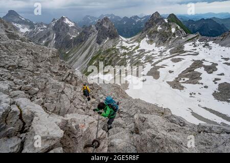 Escursionista e escursionista discesa terreno roccioso, percorso fissato con corda d'acciaio, sullo sfondo panorama montano con vecchi campi di neve e montagna rocciosa Foto Stock