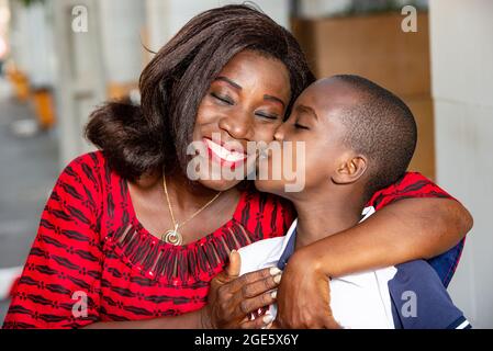 Bella donna africana in tovaglia rossa e bel bambino vestito di bianco blu fa un bel bacio sulla guancia sinistra di sua madre Foto Stock