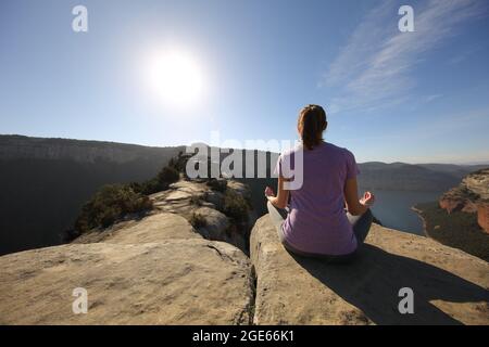 Indietro ritratto di una persona che fa esercizio di yoga in cima a una scogliera in montagna di fronte al sole Foto Stock