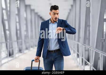 Tardi per il volo. Ritratto dell'uomo arabo preoccupato che controlla il tempo sull'orologio da polso mentre cammina con la valigia al terminal dell'aeroporto, Mal Medio Oriente emotivo Foto Stock