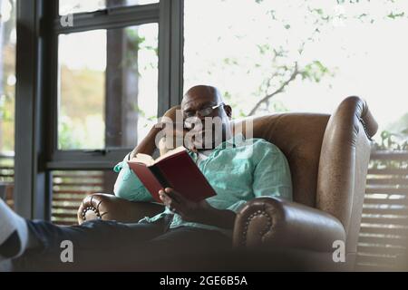 Uomo africano americano anziano seduto sulla poltrona e libro di lettura nel soggiorno moderno Foto Stock