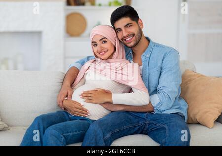 Amando invitare la coppia musulmana cuddling mentre trascorre il tempo insieme a casa, sorridente alla macchina fotografica, spazio libero. Una coppia araba incinta seduta sul divano al l Foto Stock