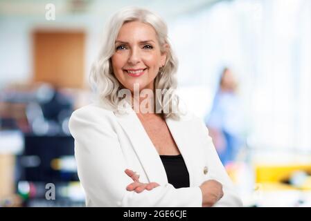 Ritratto di bella donna d'affari anziana con capelli grigi in piedi in ufficio mentre si guarda la macchina fotografica e sorridente. Foto Stock