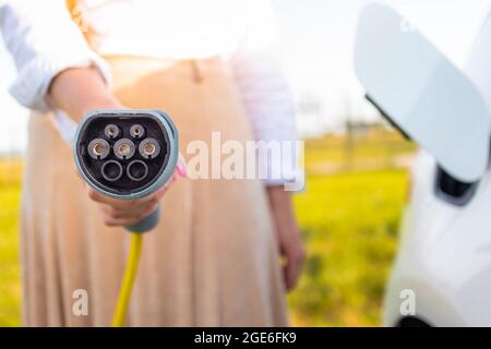 Primo piano di una mano femminile che tiene un caricatore elettrico per auto sullo sfondo visibile auto e centrale elettrica Foto Stock