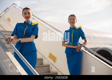 Due bellissime camere d'aria in uniforme blu che sorridono alla telecamera, in piedi su un'aerostazione e accogliendo i passeggeri Foto Stock