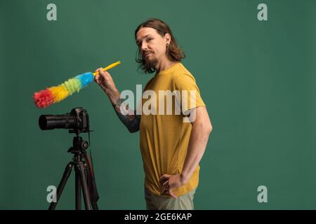 Ritratto comico di giovane uomo, fotografo, cameraman con fotocamera retrò divertimento isolato su sfondo verde studio. Concetto di occupazione, lavoro Foto Stock