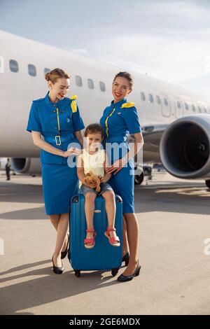 Foto a lunghezza intera di sorridenti belle stewardesses in piedi vicino cute bambina seduta sulla valigia di fronte al grande aereo e sorridente alla macchina fotografica Foto Stock