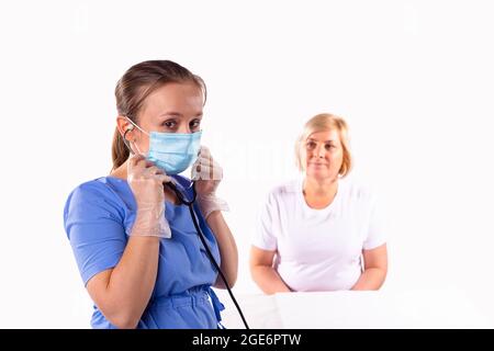 Il medico donna in uniforme, maschera medica e guanti protettivi si inza su uno stetoscopio per ascoltare la respirazione del paziente. Foto Stock