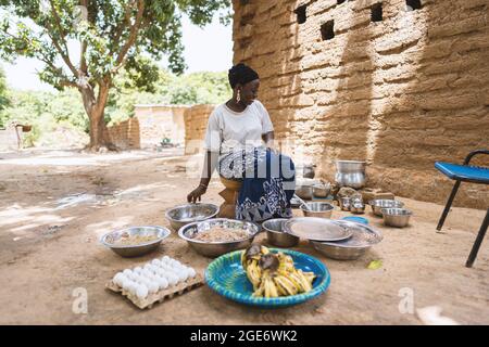 Contenuto giovane casalinga africana seduta nel mezzo di pentole e padelle nella sua cucina rustica all'aperto cucinando un pasto Foto Stock
