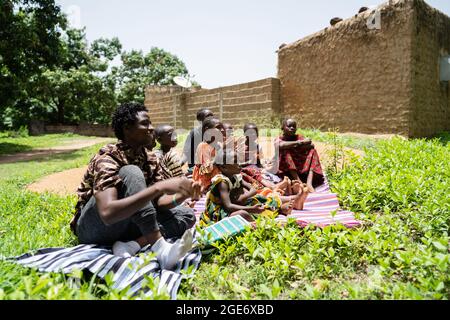 Gruppo di bambini seduti in erba che ascoltano qualcuno durante le lezioni estive in Africa occidentale Foto Stock