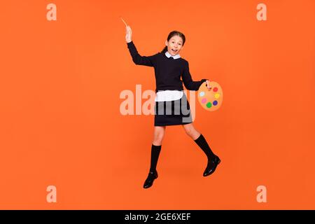 Foto di pittore carino schoolgirl salto tenere spazzola tavolozza indossare uniforme isolato arancione colore sfondo Foto Stock