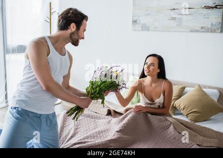 uomo allegro che tiene bouquet di fiori vicino alla fidanzata sdraiata sul letto Foto Stock