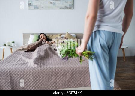 uomo offuscato che tiene bouquet di fiori vicino girlfriend sdraiato sul letto Foto Stock