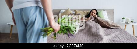 uomo che tiene bouquet di fiori vicino girlfriend sdraiato su letto, banner Foto Stock