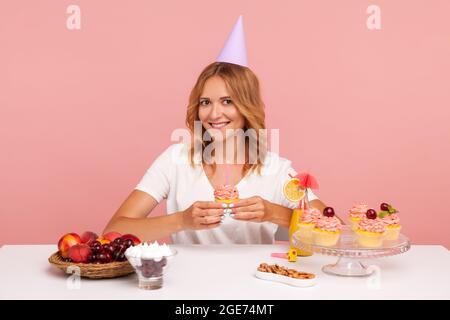 Ritratto di sorridente bella donna bionda si siede a tavola con deliziosi dessert e frutta, tenendo la torta con candela, guardando la macchina fotografica con lo felice Foto Stock