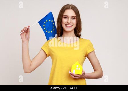 Sorridente teen ragazza agenzia immobiliare lavoratore in giallo T-shirt che tiene carta giocattolo casa e bandiera di unione europea, guardando la macchina fotografica con sorriso toothy. IO Foto Stock