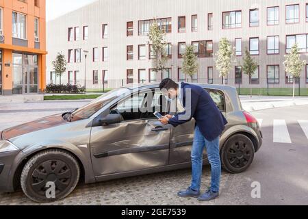 Agente assicurativo che scatta foto di auto distrutte, bruna maschio indossando maschera medica chirurgica, jeans e giacca vicino a auto schiantata sulla strada della città, Foto Stock