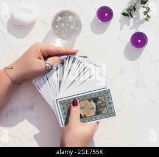 Mani con unghie viola e anelli tenere mazzo di carte Tarocchi su superficie bianca con palla di cristallo, candele e pietre. Vista dall'alto Foto Stock