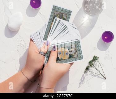 Mani con unghie viola e anelli tenere mazzo di carte Tarocchi su superficie bianca con palla di cristallo, candele e pietre. Vista dall'alto Foto Stock
