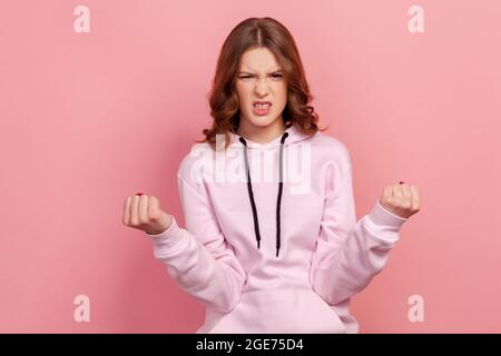 Ritratto di furiosa femmina adolescente in piedi felpa con cappuccio con denti aggrappati e mani rialzate che esprimono rabbia selvaggia, sentendosi pazzo. Studio interno sh Foto Stock