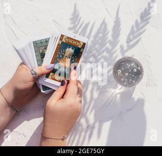 Mani con unghie viola e anelli tenere il mazzo di carte Tarocchi su superficie bianca con palla di cristallo e l'ombra foglia di felce. Vista dall'alto Foto Stock