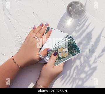 Mani con unghie viola e anelli tenere il mazzo di carte Tarocchi su superficie bianca con palla di cristallo e l'ombra foglia di felce. Vista dall'alto Foto Stock