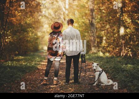 Elegante coppia in piedi con il cane carino in soleggiati boschi autunnali. Escursione in famiglia con il cane bianco del pastore svizzero. Viaggiare con animali domestici, fedele com Foto Stock