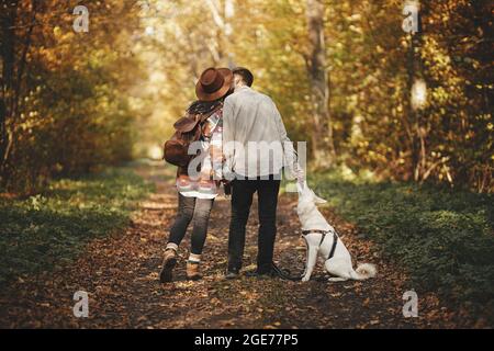 Elegante coppia baciante e in piedi con il cane carino in soleggiati boschi autunnali. Escursione in famiglia con il cane bianco del pastore svizzero. Viaggiare con pe Foto Stock