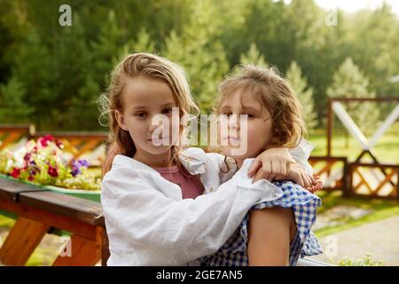 Adorabile ragazza in camicia bianca abbracciando l'amico in abito a scacchi e guardando la macchina fotografica mentre si riposa vicino alla ringhiera terrazza il giorno del fine settimana estivo Foto Stock