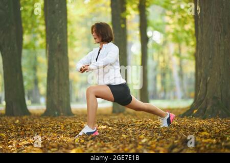 Felice donna attiva in outfit sportivo stretching corpo al parco locale durante l'esercizio mattutino. Giovane donna con taglio corto che conduce uno stile di vita sano. Attività all'aperto. Foto Stock