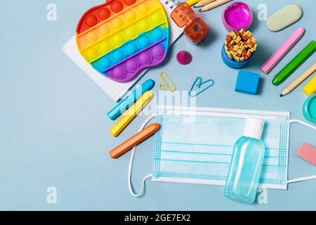 Forniture scolastiche, antipasto, maschera facciale, bottiglia di disinfettante, per il ritorno a scuola su sfondo blu. Precauzioni Covid-19. Vista dall'alto in piano Foto Stock