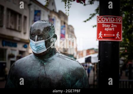 'The Sockman' dell'artista scozzese Shona Kinloch rappresenta l'industria calzetteria di Loughborough e qui visto con una maschera facciale accanto a un durin segno Covid Foto Stock