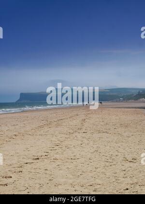 Un'enorme distesa di spiaggia vuota e sabbiosa con impronte che conducono ad un gruppo lontano di persone in silhouette e Hunt Cliff, il tutto sotto un cielo azzurro e nocciola. Foto Stock
