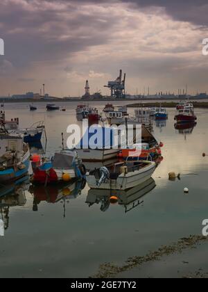 Piccole barche da pesca a riposo in acque cristalline davanti ad un orizzonte industriale, sotto un cielo ostico. Foto Stock