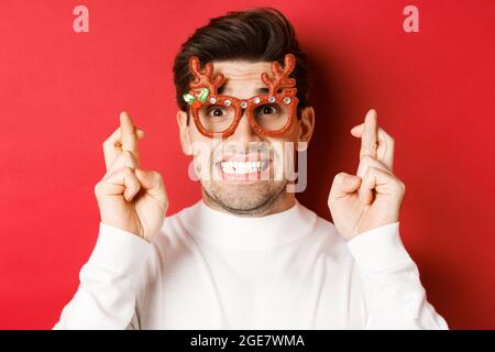 Concetto di vacanze invernali, natale e celebrazione. Primo piano di uomo speranzoso in occhiali da festa, incrociando le dita e facendo un desiderio per il nuovo anno, stand Foto Stock