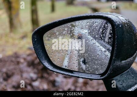 Primo piano dello specchietto retrovisore laterale della vettura con gocce di pioggia su sfondo sfocato. Giornata piovosa sulla strada. Guida in cattive condizioni meteorologiche. Foto Stock