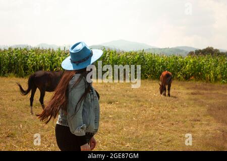 stile di vita rurale donna con capelli rossi che cammina attraverso i campi di mais indossando giacca e cappello in denim per proteggersi dal sole guardando le piante an Foto Stock