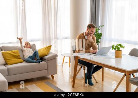 Bianco uomo caucasico che lavora con il suo portatile mentre suo figlio sta giocando sul divano Foto Stock