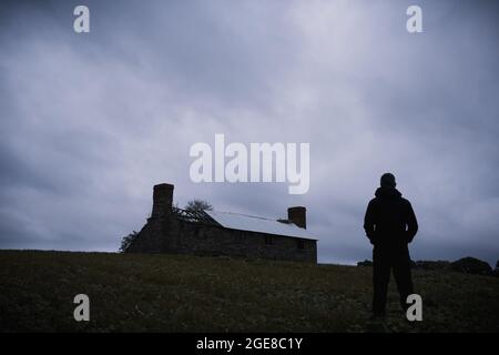 Un angolo basso, di una misteriosa figura incappucciata, guardando una casa rotta e abbandonata nel mezzo della campagna. In una serata di moody. Foto Stock