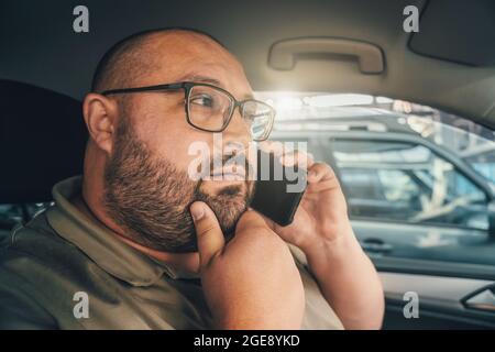 Uomo perplesso con occhiali che parlano al telefono mentre si guida in auto. Emozione di dubbio e diffidenza sul volto di divertente uomo grasso. Foto Stock