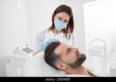 Estetista con maschera fa l'iniezione al paziente dell'uomo durante la procedura di terapia meso anti-aging in clinica Foto Stock