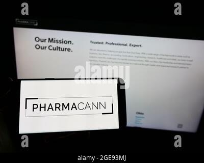 Persona che tiene il cellulare con il logo della società americana di cannabis PharmaCann Inc. Sullo schermo di fronte al sito web aziendale. Mettere a fuoco sul display del telefono. Foto Stock