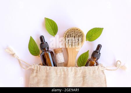 Borsa riutilizzabile in lino eco-compatibile con le diverse bottiglie e accessori da bagno in legno naturale e vetro marrone. Foglie naturali fresche intorno.C Foto Stock