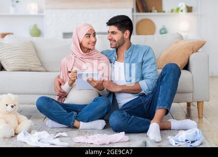Giovane coppia islamica incinta che si prepara per il parto, facendo una lista di controllo delle necessità mentre si siede a terra Foto Stock