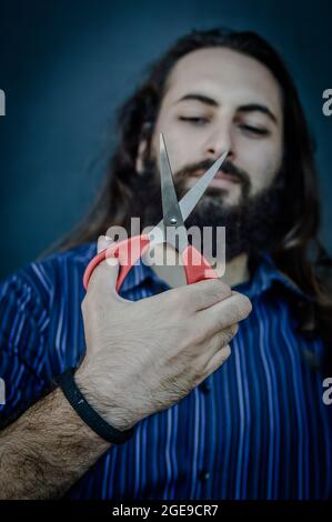 ritratto di un giovane uomo con una barba e capelli lunghi che tiene una forbice in mano e la guarda mentre pensa di tagliare tutto per cambiare Foto Stock